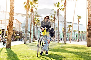 Pretty brunette girl with a bike on grass suround palms. She is chilling in sunlight, smiling to camera