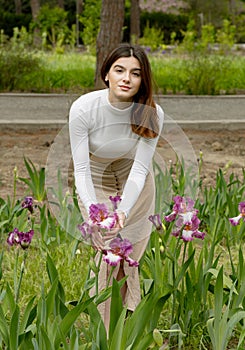 Pretty brunette girl posing in garden, bending, holding flower by hands, looking at camera.