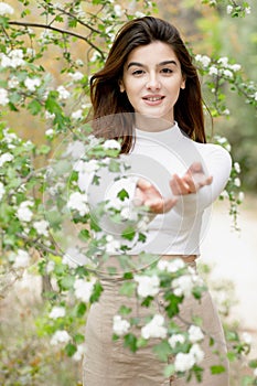 Pretty, brunette girl standing in blooming tree, stretching hands, looking at camera.