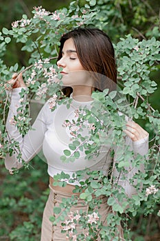 Pretty brunette girl standing, holding blooming tree branch, smelling with closed eyes.