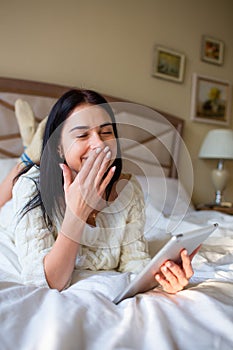 Pretty brunette girl in white knitted dress lying on bed with a tab in and hands and laughing covering her mouth with a hand