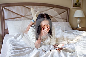 Pretty brunette girl in white knitted dress lying on bed with a tab in and hands and laughing covering her mouth with a hand