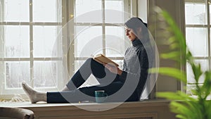 Pretty brunette in sweater and jeans is reading book sitting on window-sill on Christmas day enjoying soliture, free