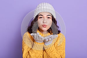 Pretty brunette woman dresses warm yellow sweater, white capand gloves sending air kiss at camera isolated over lilac background,