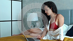 Pretty brunette woman lying on bed in apartment and texting on computer. Cheerful smiling attractive female in lingerie