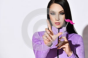 Pretty brunette woman in purple clothes gestures with finger and holding decorative pink mustache in hand