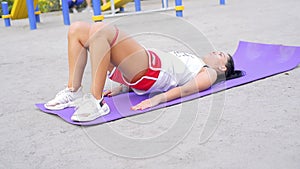 Pretty brunette woman train her gluteal muscles on the purple karemat at warm summer day at the park