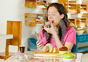 Pretty brunette woman wearing pink shirt sitting at table inside bakery, biting into slice of bread