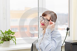 pretty business woman in glasses and a blue blouse stands at the desktop in the office against the background of a light window