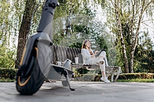 Pretty business woman in a white suit sits in a city park, drinks coffee and works on a digital tablet with an electric scooter in