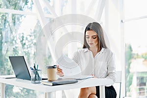 Pretty businesswoman sitting at workplace and reading documents in her modern office