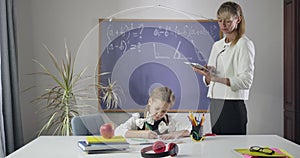 Pretty Caucasian girl drawing in exercise book as adult private teacher searching tasks in tablet. Child studying at