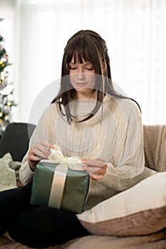 A pretty Caucasian girl is unwrapping a Christmas gift on a couch in her living room on Christmas