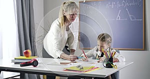 Pretty caucasian schoolgirl writing with red pencil while adult teacher talking to her. Cute child studying at home with