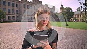 Pretty caucasian short-haired blonde woman is using her tablet and standing still on the sunndy urban street