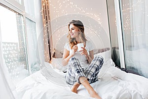 Pretty caucasian woman in cozy attire sitting at loggia. Indoor photo of curious female model in white t-shirt drinking