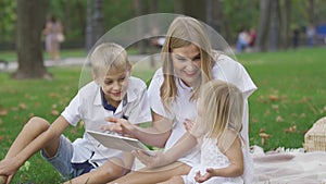 Pretty caucasian woman and her two kids spending time outdoors. The woman showing her children photos on her tablet