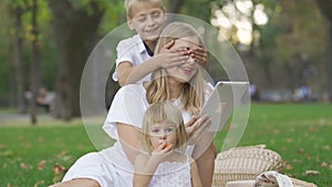 Pretty caucasian woman and her two kids spending time outdoors. The woman using her tablet, daughter sitting near, the