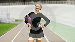 Pretty caucasian woman standing near running track and posing with fitness rug. Healthy lifestyle concept