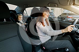 Pretty Caucasian woman, young mother driving a car, having her little baby girl in a child seat behind. Safety driving