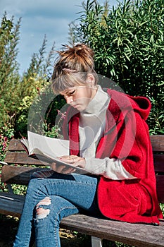Pretty Caucasian Young Girl in Red Jacket Reading a Book Sitting on a Park Bench