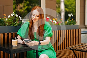 Pretty Caucasian young woman using browsing smartphone looking at screen, sitting at table with coffee cup in outdoor