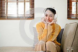 Pretty asian woman relaxing on sofa in her living room, listening to music through headphones