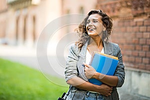 Pretty cheerful student smiling at camera carrying notebook on campus at college