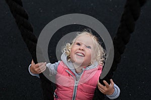 Pretty cheerful toddler girl with blond curly hair and big blue eyes happily swinging on a swing, made of black rope