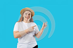 Pretty cheerful woman in straw hat gesturing with fingers and showing away isolated over blue background, studio