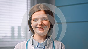 Pretty cheerful young woman general practitioner looks straight and smiles standing against blue hospital room wall