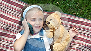 Pretty child girl laying on green lawn with her teddy bear talking on mobile phone