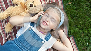 Pretty child girl laying on green lawn with her teddy bear talking on mobile phone