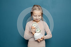 Pretty child girl putting in piggy bank one dollar banknote on blue background