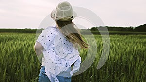 Pretty child in the hat is running across the wheat field at sunset.