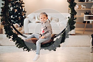 Pretty child sitting on Christmas swing. Christmas tree.