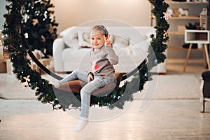 Pretty child sitting on Christmas swing. Christmas tree.