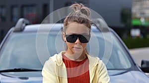 Pretty cool young teenager girl in black sunglasses standing in front of car on street looking at camera chewing a gum