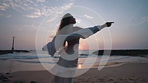 Pretty curly girl posing, long hair blowing in wind at amazing sunset on beach. Young woman enjoying summer sunrise at