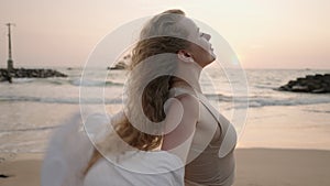 Pretty curly girl posing, long hair blowing in wind at amazing sunset on beach. Young woman enjoying summer sunrise at