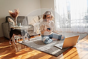 Pretty curly happy young woman sitting at home on the floor in front of laptop with her pet dog working and learning online with