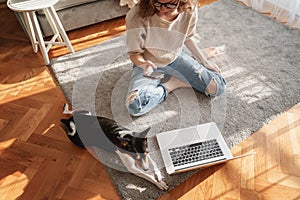 Pretty curly happy young woman sitting at home on the floor in front of laptop with her pet dog working and learning online with