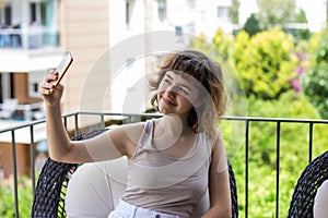 Pretty curly smiling young girl taking a selfie on smartphone on the apartment balcony. Video chatting with friends