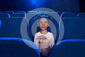 Pretty cute girl sitting with popcorn bucket in cinema.