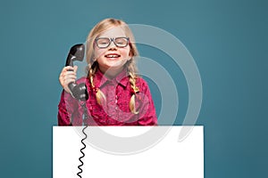 Pretty, cute little girl in pink shirt, black trousers and glasses holds empty poster and having a phone talk