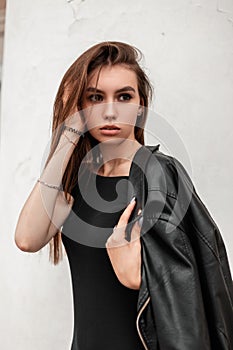Pretty cute young woman in an autumn leather black jacket in a fashionable black dress straightens hair standing near a white wall