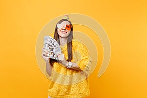 Pretty dreamy young woman in orange heart glasses birthday party hat looking up holding bundle lots of dollars cash