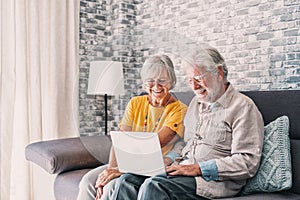 Pretty elderly 70s grey-haired couple resting on couch in living room hold on lap laptop watching movie smiling enjoy free time,