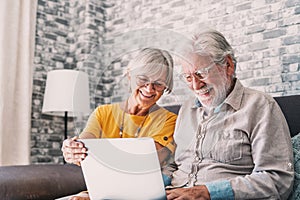 Pretty elderly 70s grey-haired couple resting on couch in living room hold on lap laptop watching movie smiling enjoy free time,