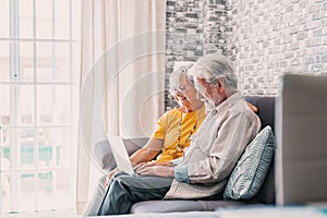 Pretty elderly 70s grey-haired couple resting on couch in living room hold on lap laptop watching movie smiling enjoy free time,